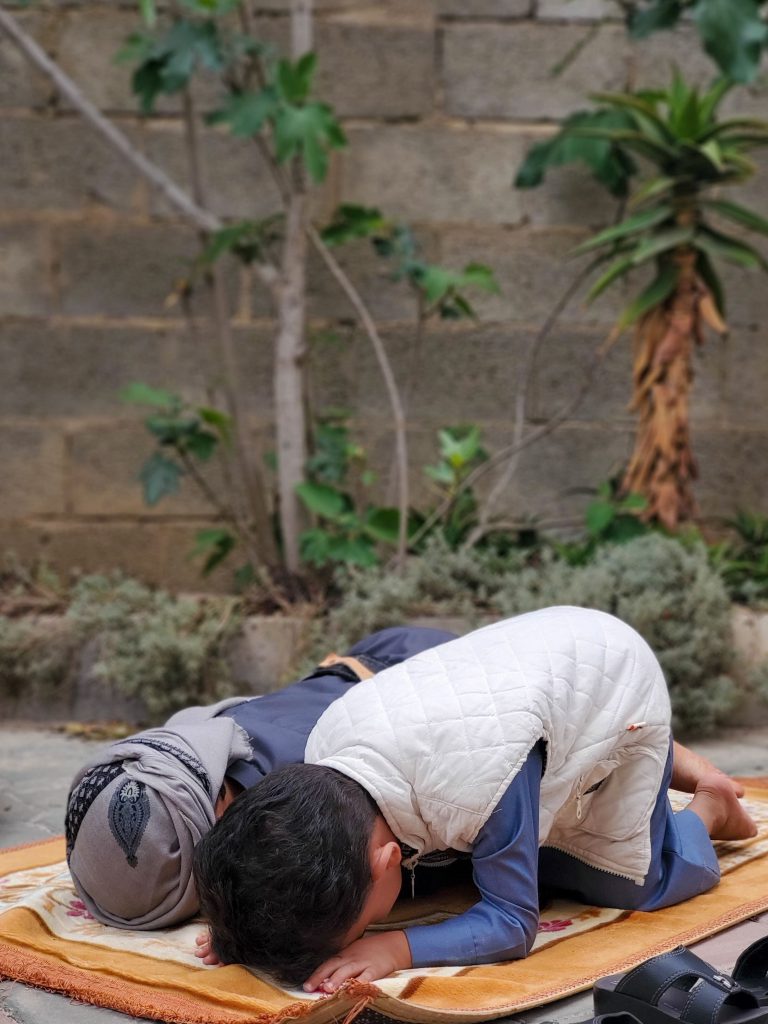 A young boy in traditional dress praying outdoors in Sanaa, Yemen, surrounded by plants.